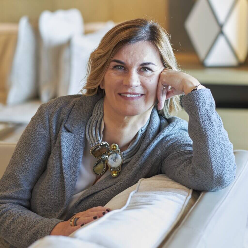 A smiling woman with blonde hair, wearing a grey jacket and an elaborate statement necklace, relaxes on a light-colored couch.