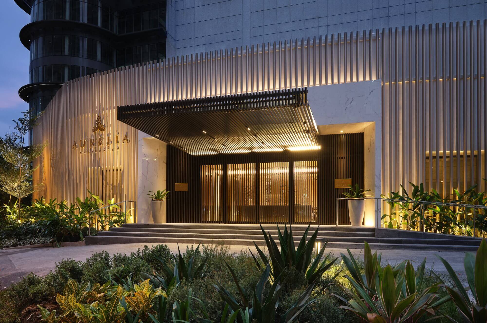 Aurelia Residences showsuite facade at dusk, featuring a modern design with vertical slats, an illuminated entrance canopy, and lush landscaping.